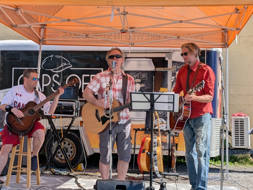 Three members of Boxcar Junction performing under an orange Caps Off Brewing tent.
